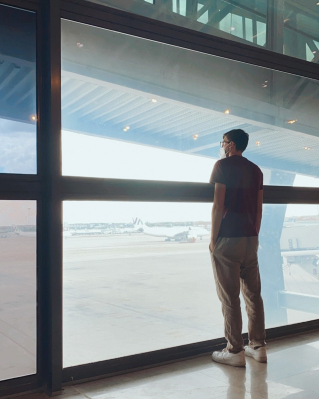 A men stands inside an airport and looking outside the window to look at the planes and flights