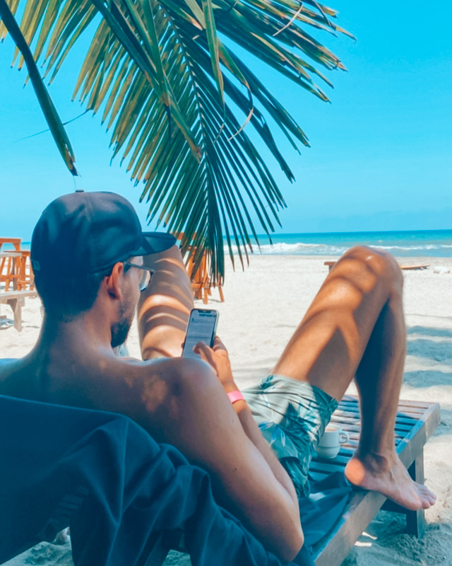 Guy sits at a beach checking his travel apps