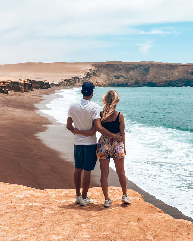 A couple is standing at a red beach, looking at the ocean and the desert in Paracas, Peru.