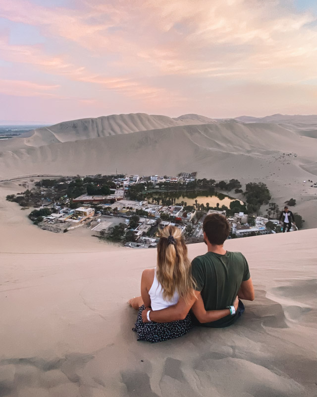 A couple sits on a high sand dune and is watching the sunset over Huacachina, Peru